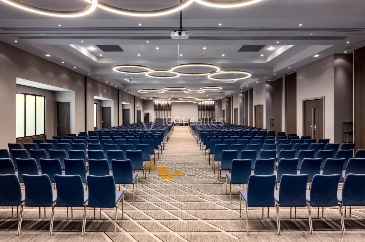 Salle de conférence moderne avec des rangées de chaises bleues alignées et des luminaires circulaires au plafond.