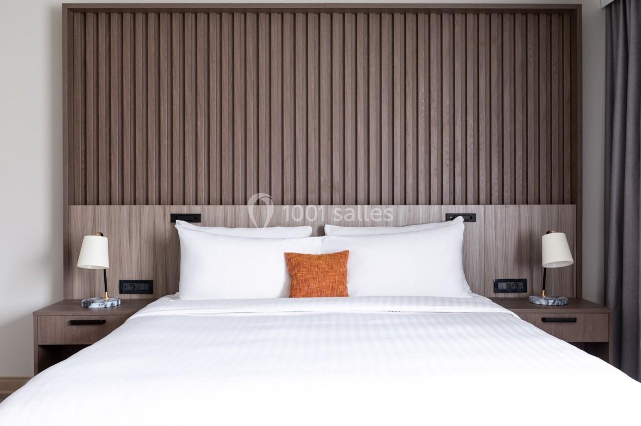 Lit double avec draps blancs, coussin orange, deux lampes de chevet et une tête de lit en bois à lattes verticales.