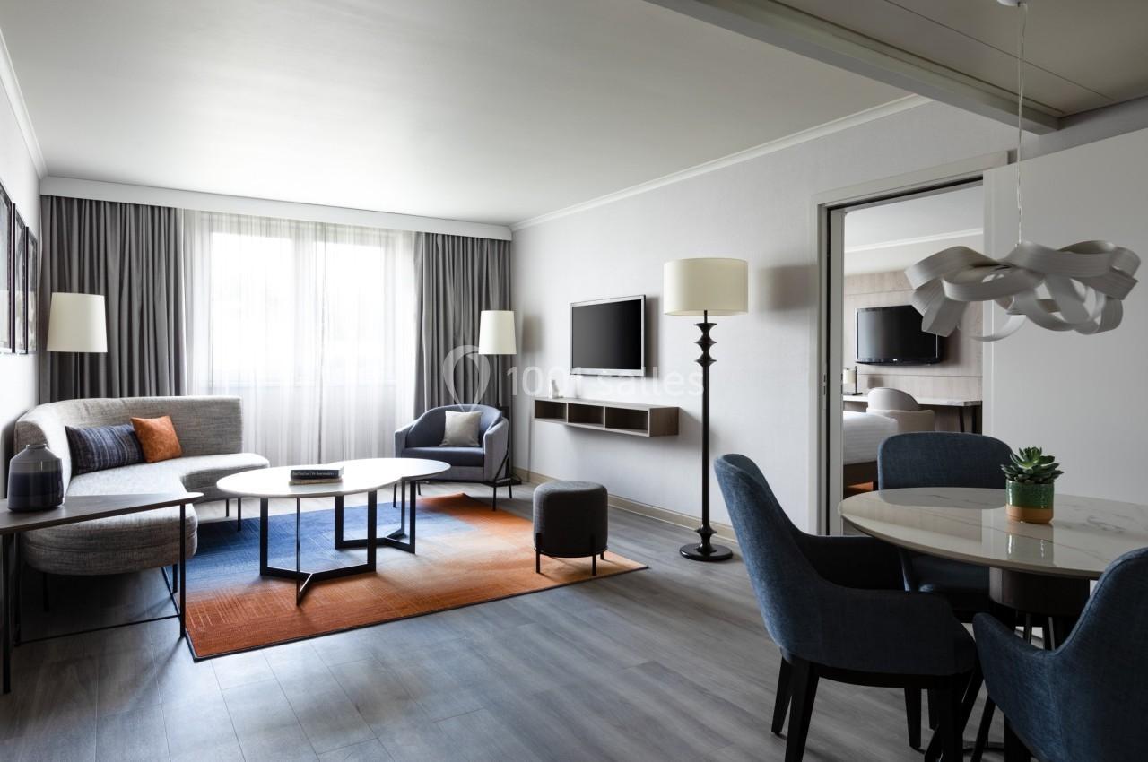 Salon moderne avec canapé, table basse, télévision murale, coin repas et grande fenêtre laissant entrer la lumière naturelle.