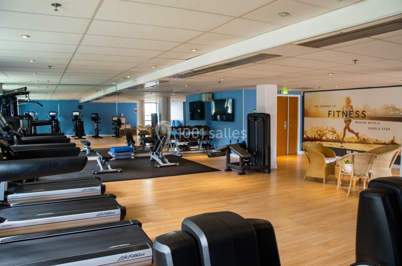 Salle de sport lumineuse avec tapis de course, vélos, haltères et coin détente avec fauteuils en osier.