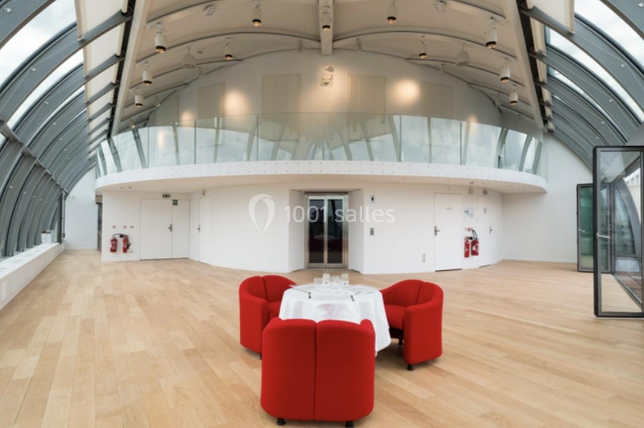 Salle lumineuse avec toit en verre courbé, parquet clair et table entourée de fauteuils rouges au centre.