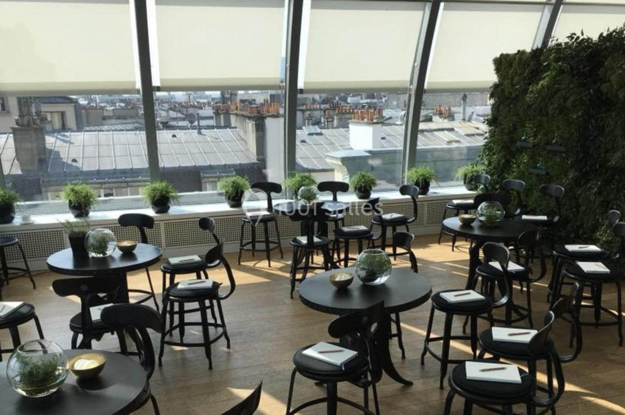 Salle lumineuse avec des tables rondes noires, des chaises assorties, des plantes et une grande baie vitrée donnant sur des…