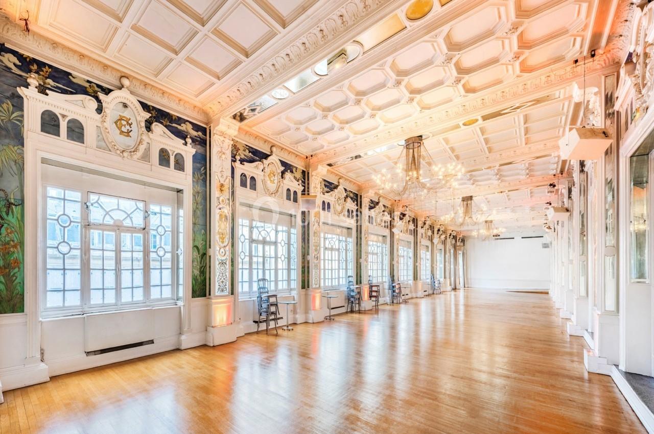 Salle lumineuse avec parquet en bois, grandes fenêtres ornées de vitraux et plafond décoratif avec moulures.