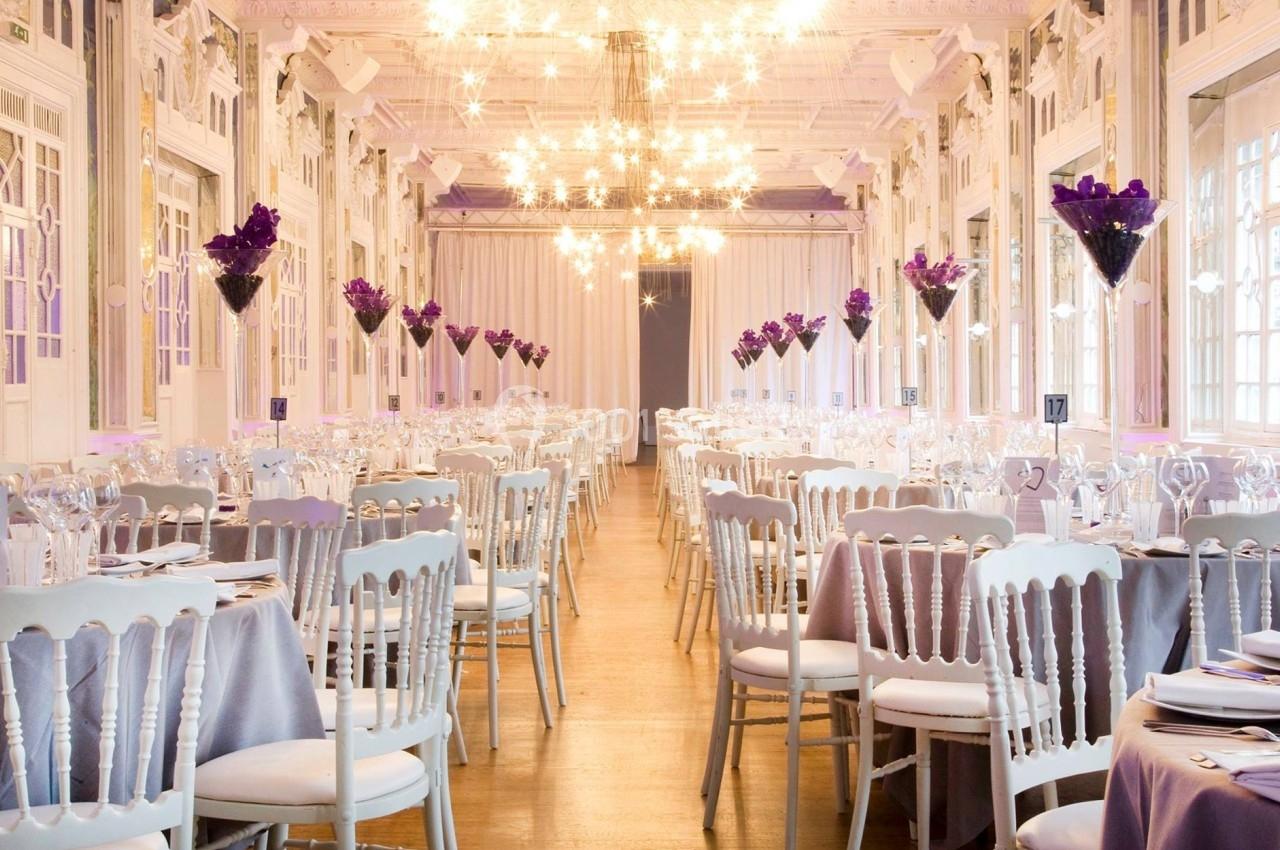 Salle de réception élégante avec tables dressées, chaises blanches et décorations florales violettes sous des lustres…