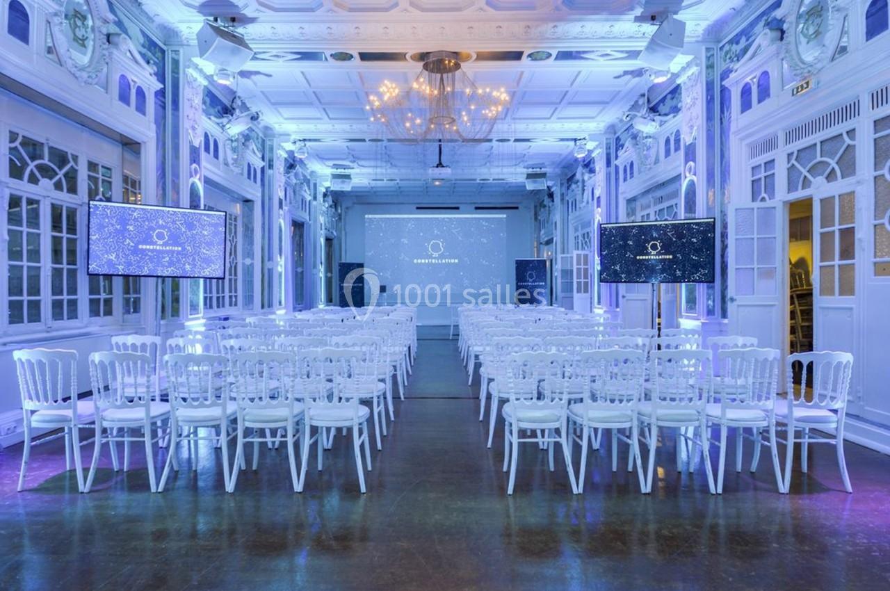 Salle de conférence lumineuse avec des rangées de chaises blanches, deux écrans et un projecteur au fond.