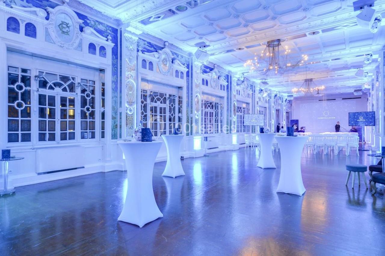 Salle de réception lumineuse avec hauts plafonds, grandes fenêtres, tables hautes et éclairage bleu.