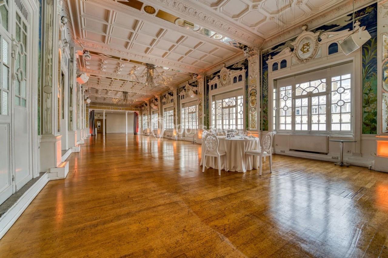 Salle élégante avec parquet en bois, grandes fenêtres, murs ornés et une table dressée dans un coin.