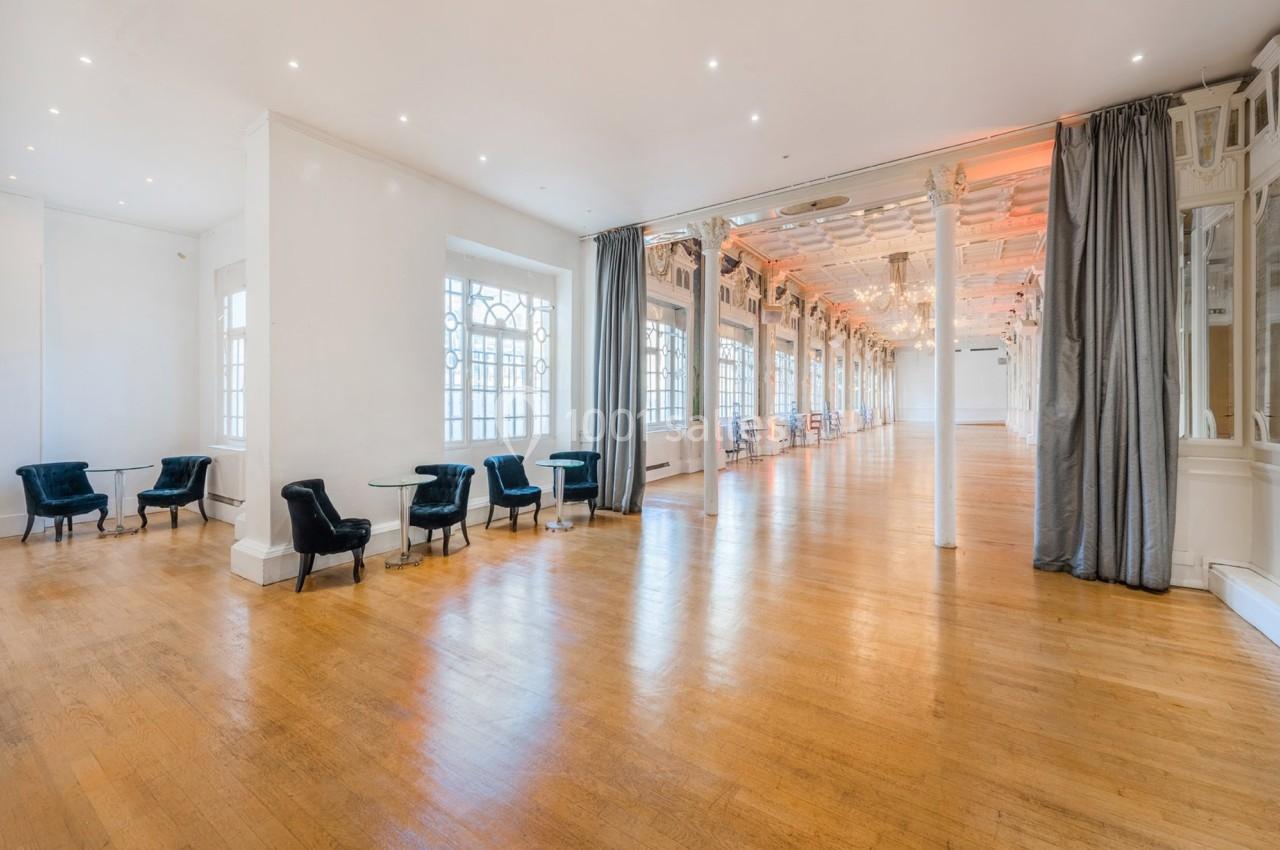Grande salle lumineuse avec parquet en bois, baies vitrées, fauteuils noirs et rideaux gris, décorée de lustres.
