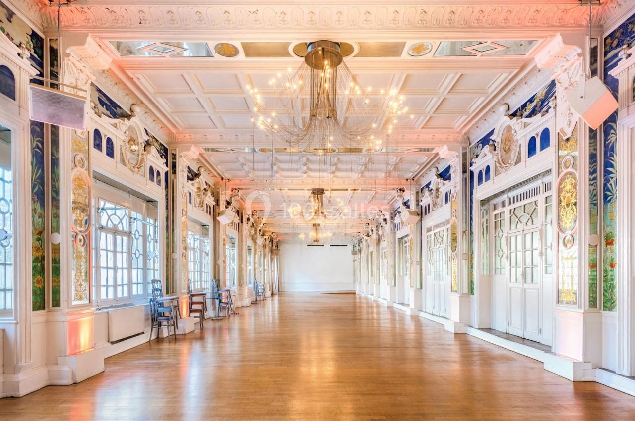 Salle spacieuse et lumineuse avec parquet, lustre central, murs ornés de fresques et grandes fenêtres.