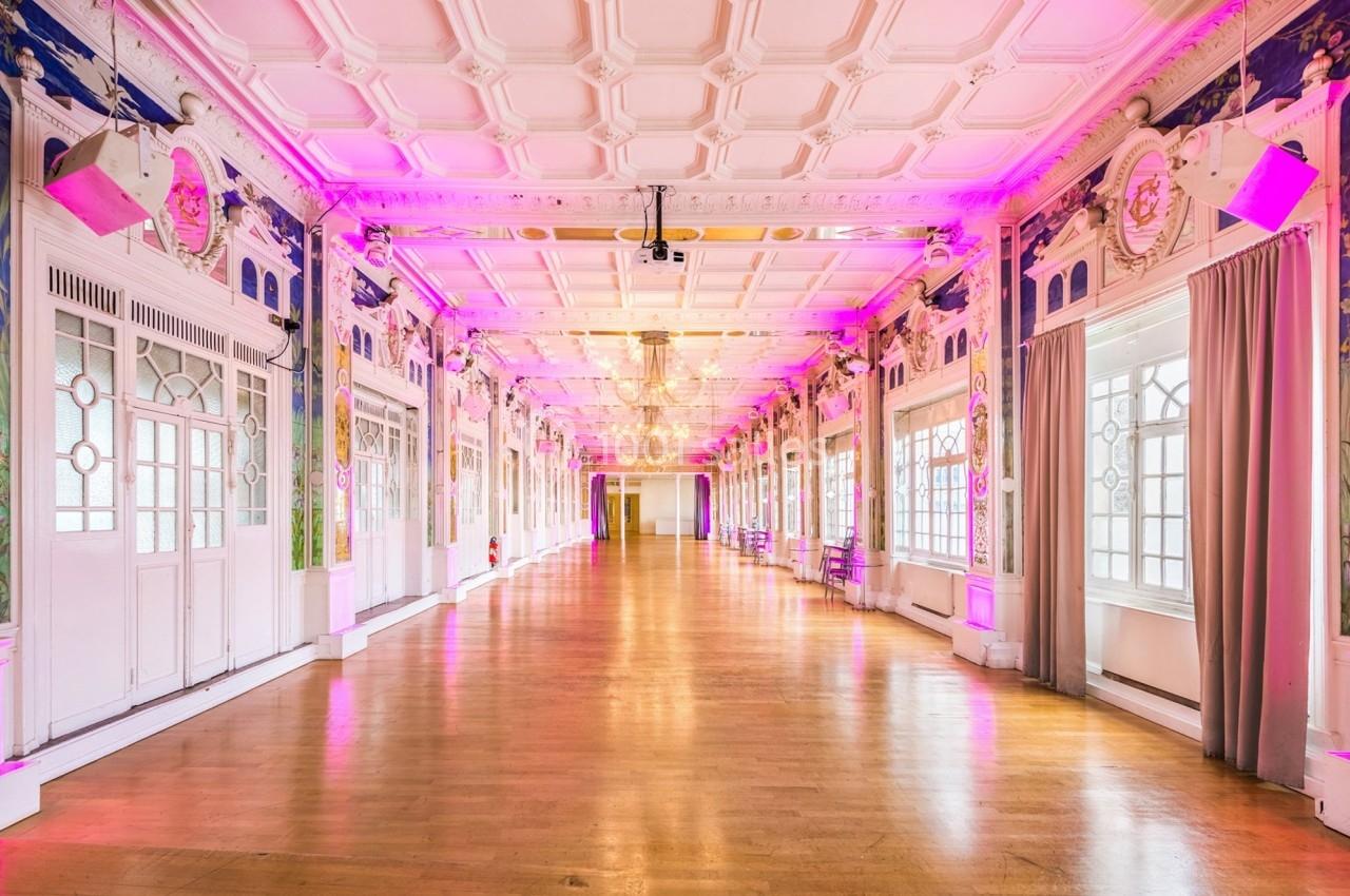 Salle spacieuse avec parquet, murs ornés et éclairage rose, grandes fenêtres laissant entrer la lumière naturelle.