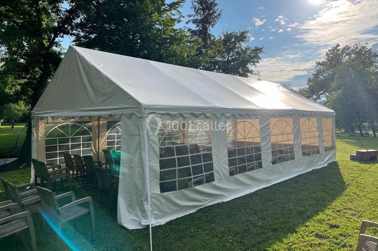 Tente blanche installée sur une pelouse ensoleillée, entourée de chaises et d'arbres.