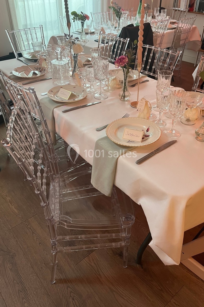 Table dressée avec nappes blanches, chaises transparentes, vaisselle élégante, fleurs et bougies dans un cadre intérieur.