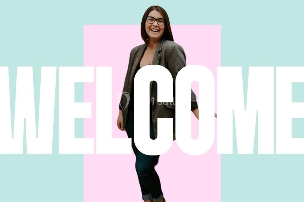 Femme souriante en blazer gris, debout devant un fond coloré avec le mot ’WELCOME’ en grandes lettres blanches.