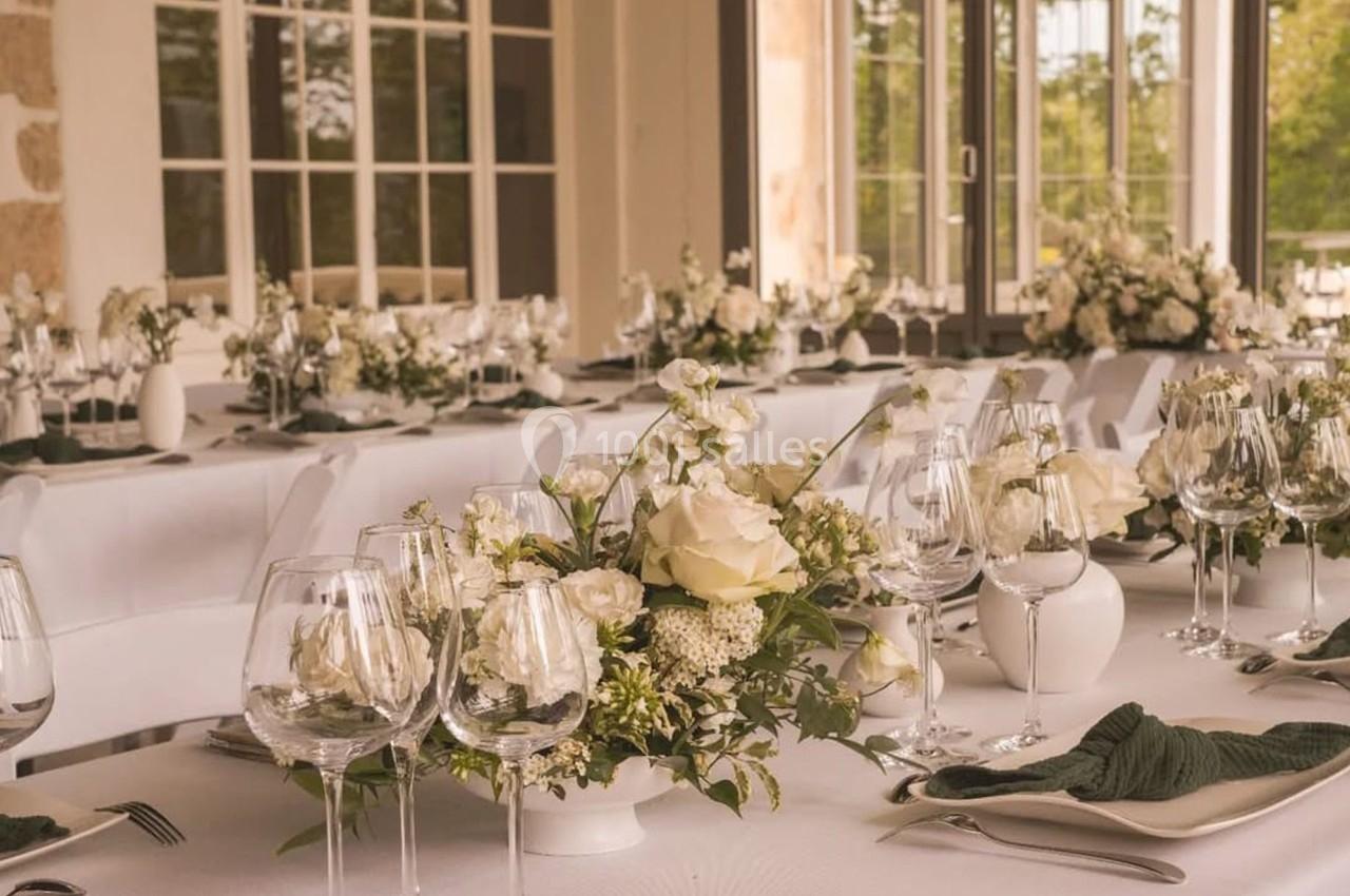 Tables élégamment dressées avec nappes blanches, compositions florales et verres à pied dans une salle lumineuse.