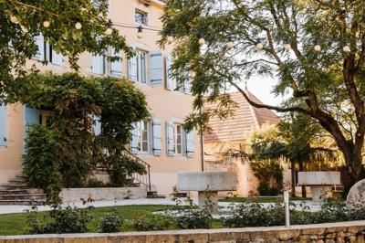 Façade d'une maison ancienne avec volets bleus, entourée de végétation et éclairée par des guirlandes lumineuses.
