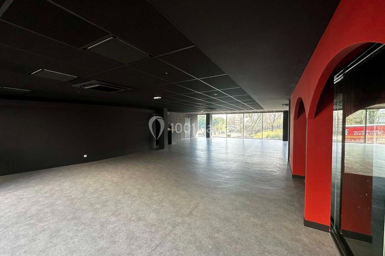 Salle vide avec sol gris, murs noirs et rouges, grandes baies vitrées laissant entrer la lumière naturelle.