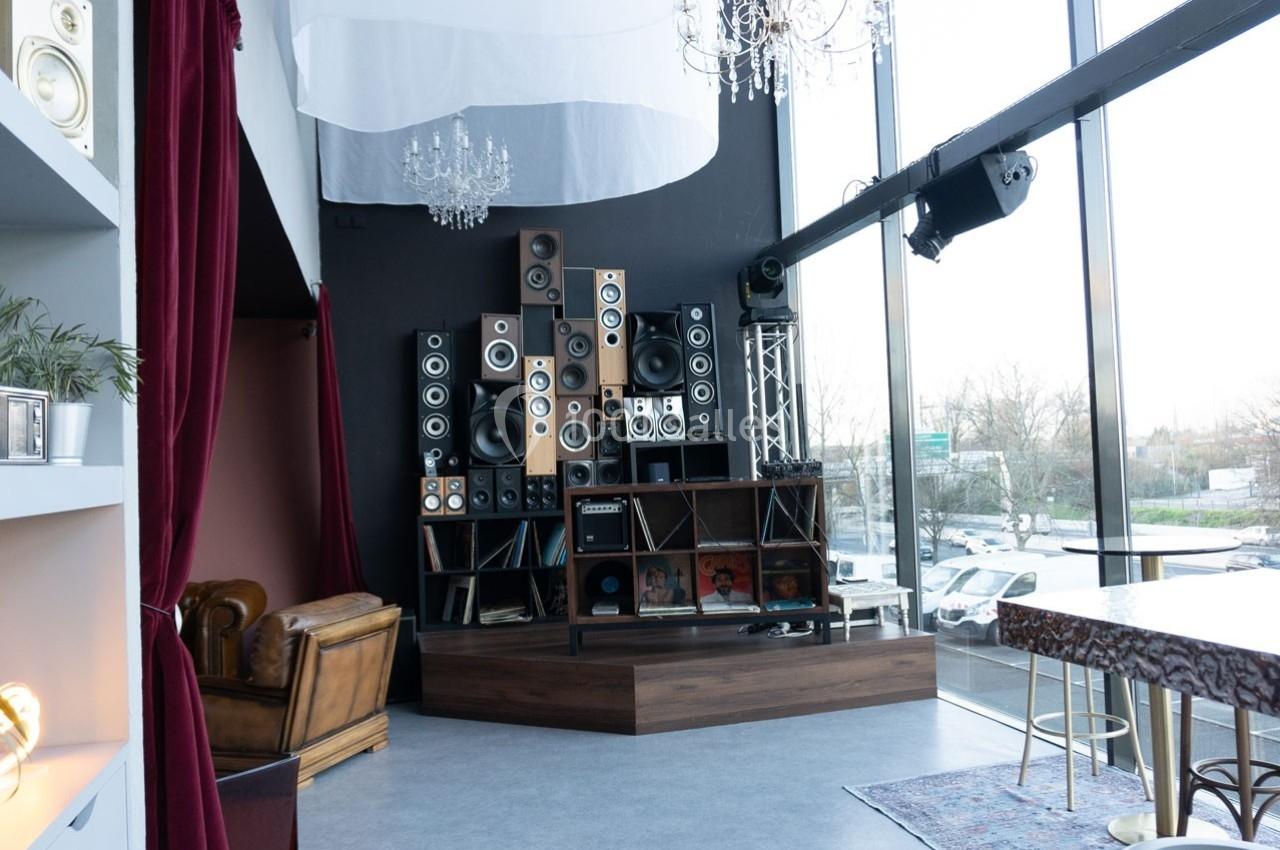 Mur de haut-parleurs empilés dans un espace lumineux avec fauteuil en cuir, rideaux rouges et grandes baies vitrées.