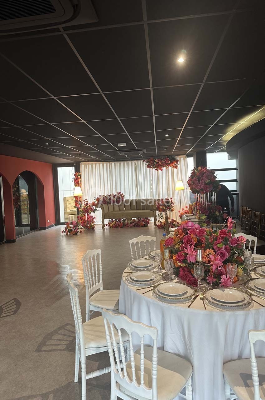 Salle décorée pour un événement avec table dressée, chaises blanches, fleurs colorées et canapé en arrière-plan.