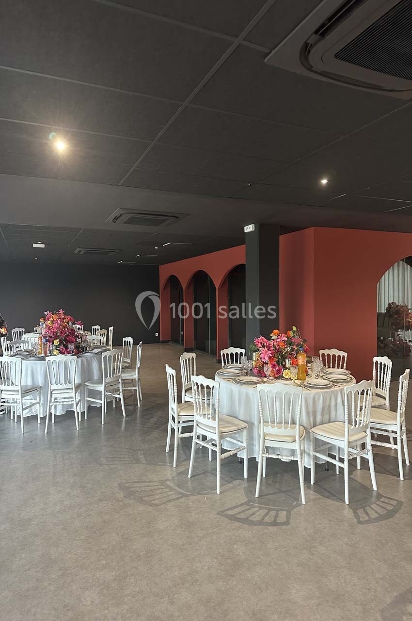 Salle de réception avec tables rondes dressées, chaises blanches et décorations florales colorées.