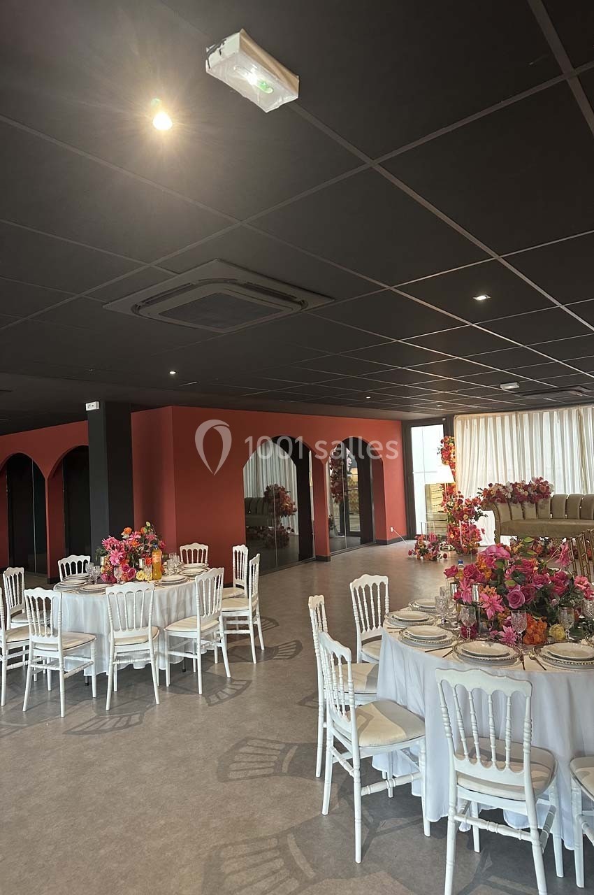 Salle de réception moderne avec tables rondes dressées, chaises blanches et décorations florales colorées.