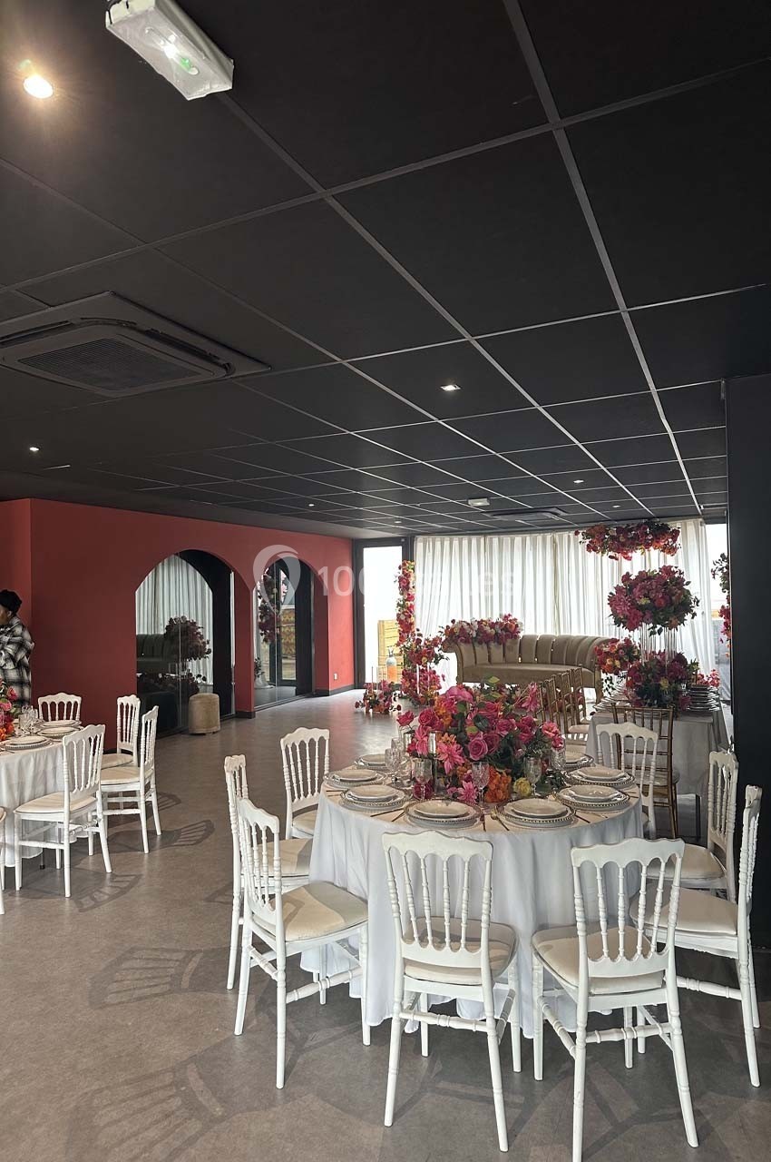 Salle de réception décorée avec des tables rondes, nappes blanches, chaises blanches et arrangements floraux colorés.