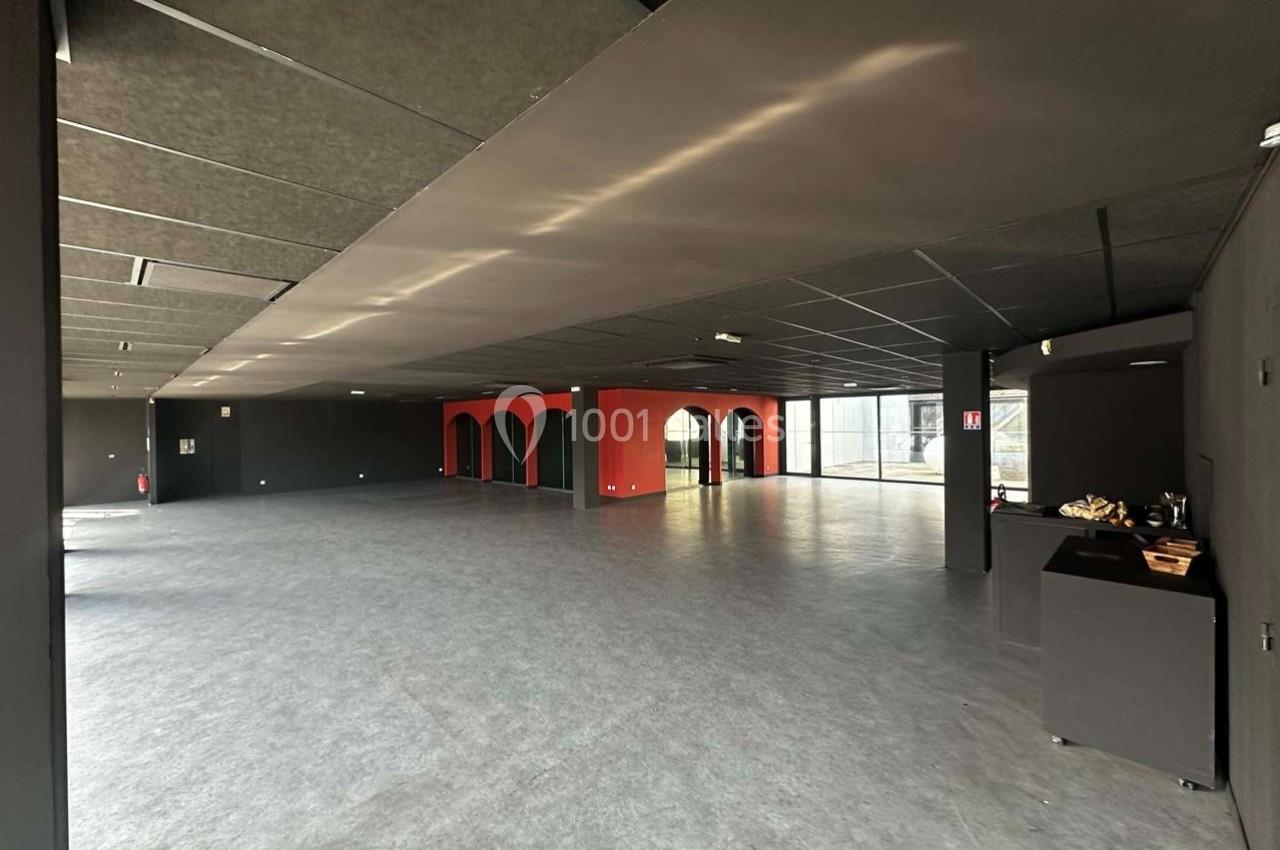 Intérieur d'un grand espace vide avec sol gris, murs sombres, arches rouges et lumière naturelle entrant par une baie vitrée.