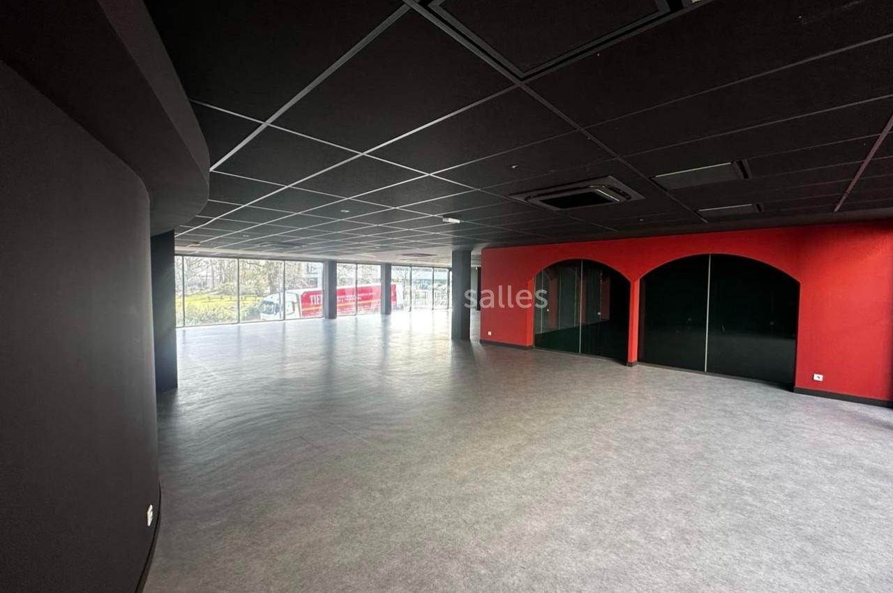 Salle vide avec sol gris, murs noirs et rouges, grandes baies vitrées laissant entrer la lumière naturelle.