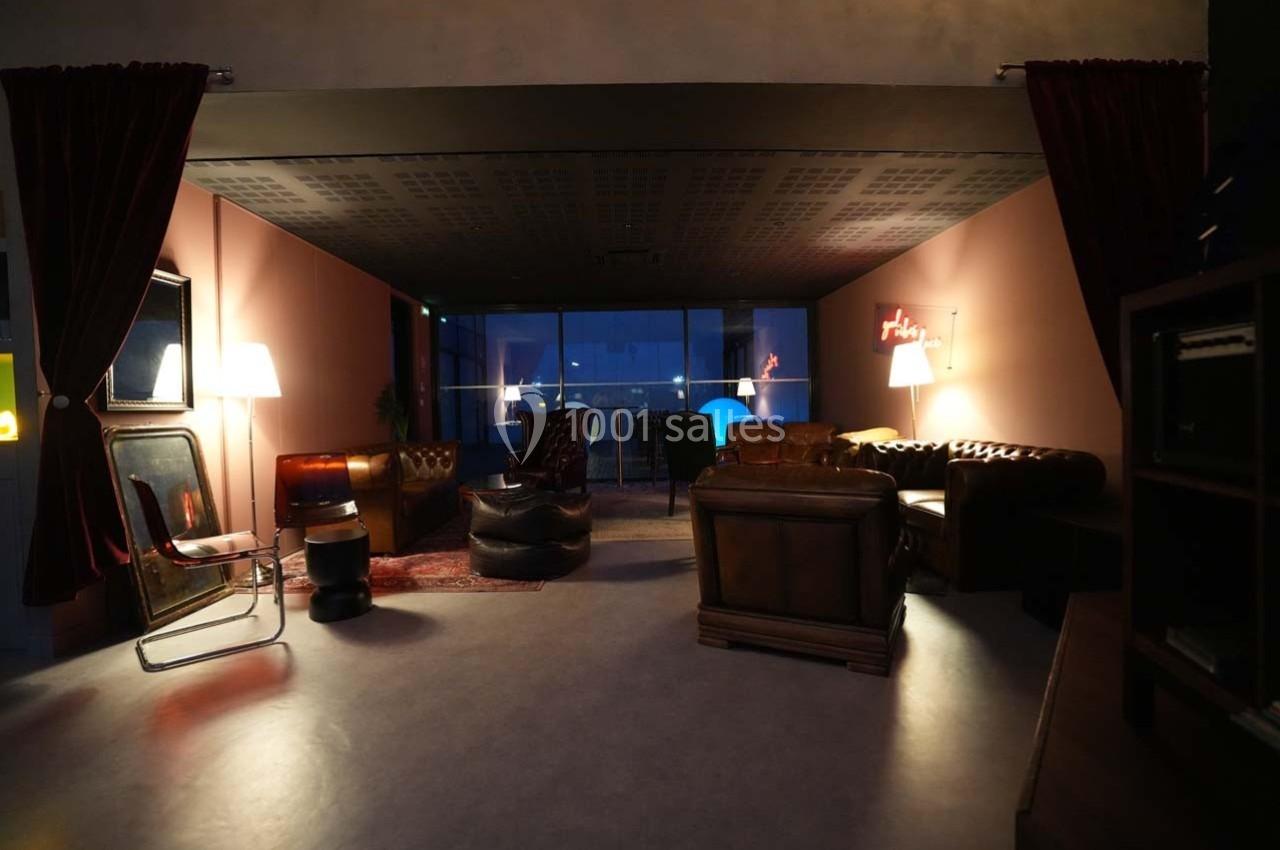 Salon cosy avec fauteuils en cuir, tapis rouge, lampes allumées et vue nocturne sur une grande baie vitrée.