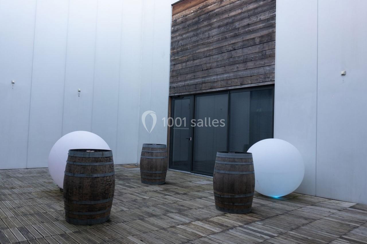 Cour intérieure avec sol en bois, quatre tonneaux et deux grandes sphères blanches près d'un mur et d'une porte vitrée.