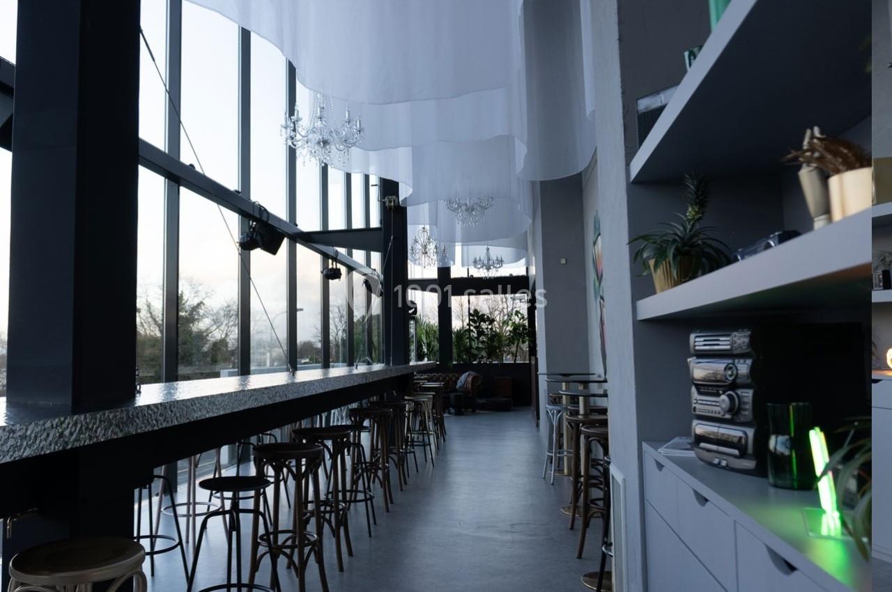 Intérieur d'un café moderne avec de grandes baies vitrées, un long comptoir, des tabourets et des étagères décorées.