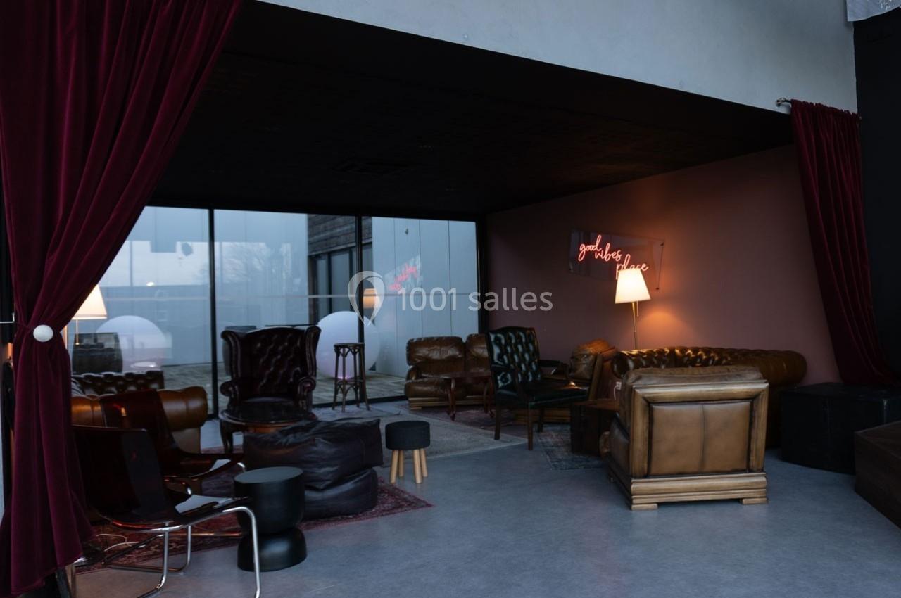 Salon cosy avec fauteuils en cuir, rideaux rouges, lampadaire allumé et enseigne néon sur un mur sombre.