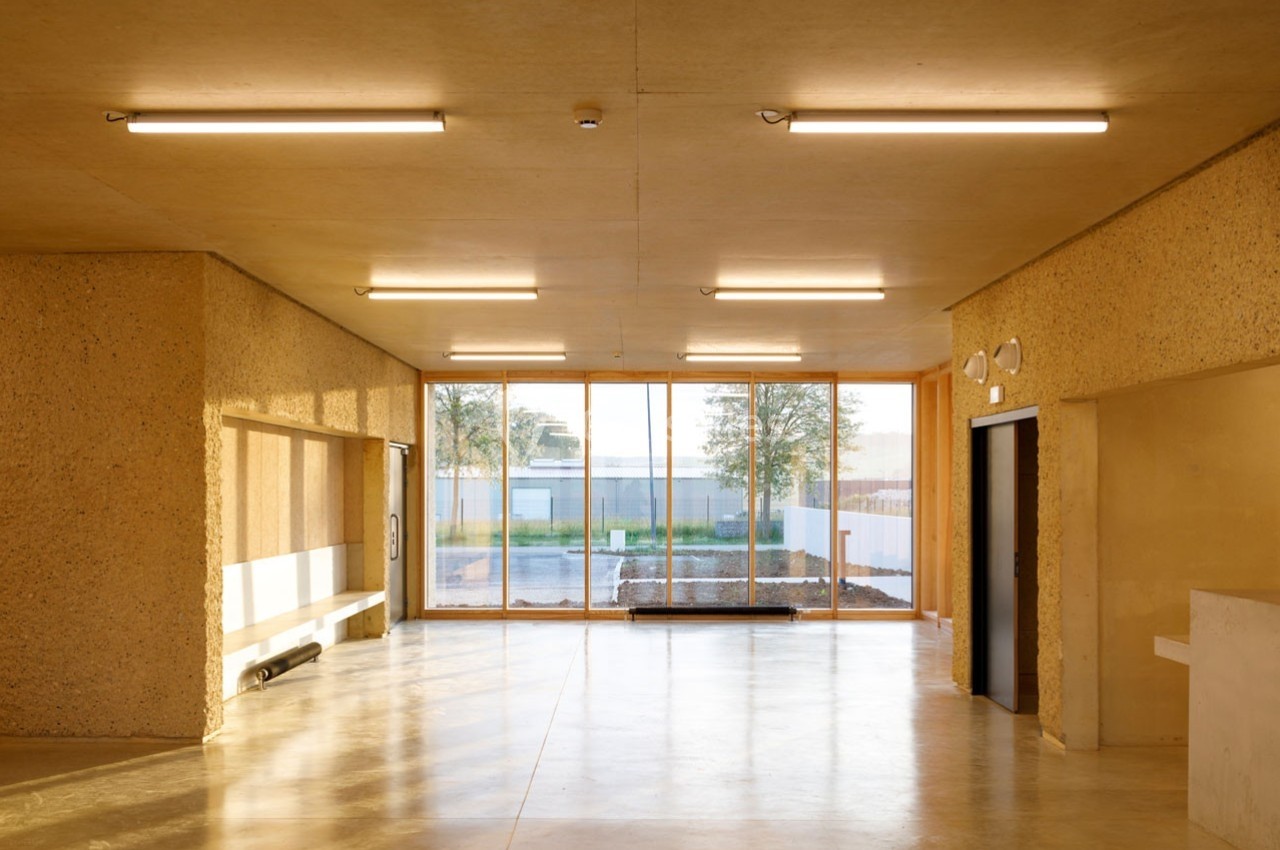 Intérieur d'un bâtiment avec murs en béton brut, sol en béton poli et grandes baies vitrées donnant sur l'extérieur.
