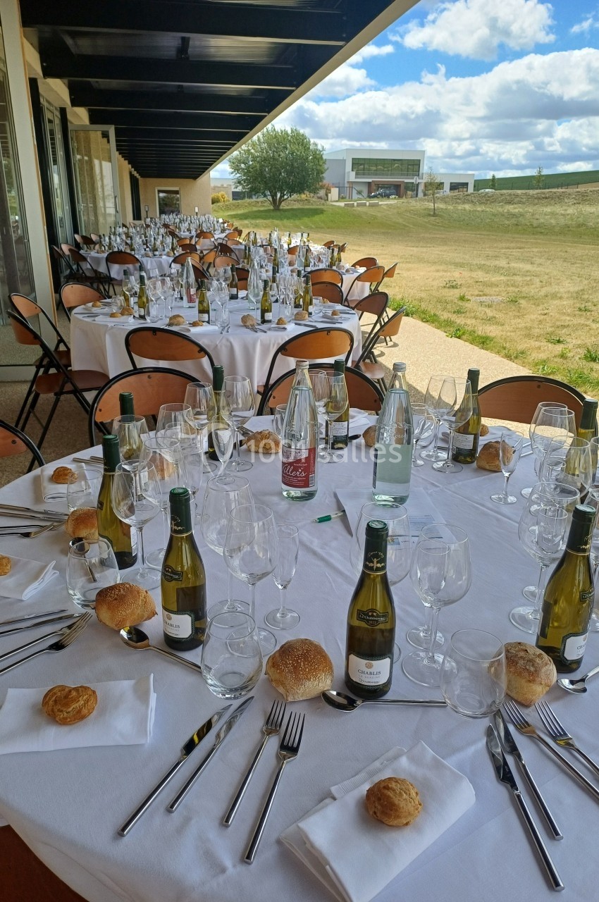 Tables rondes dressées en extérieur avec nappes blanches, bouteilles de vin, verres et pains individuels.