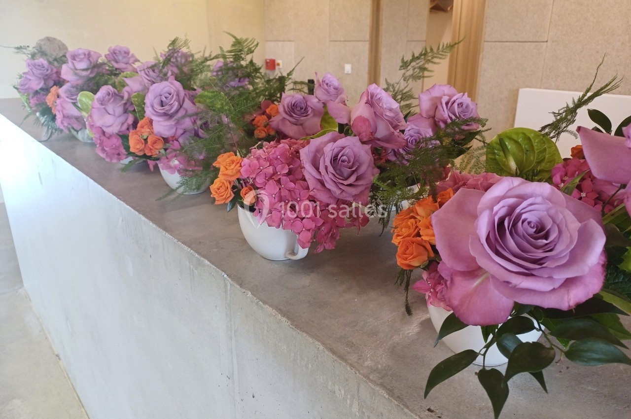 Bouquets de roses violettes, hortensias roses et petites fleurs orange alignés sur un comptoir en béton.