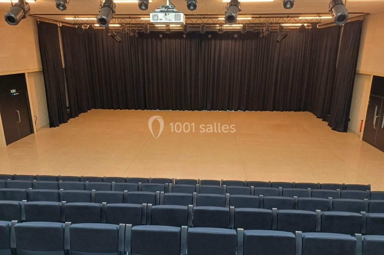 Salle de spectacle vide avec scène, rideaux noirs, projecteurs suspendus et rangées de sièges au premier plan.