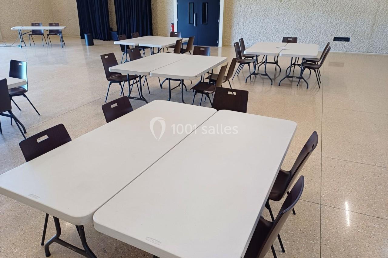 Salle avec des tables rectangulaires blanches et des chaises marron disposées sur un sol en terrazzo clair.