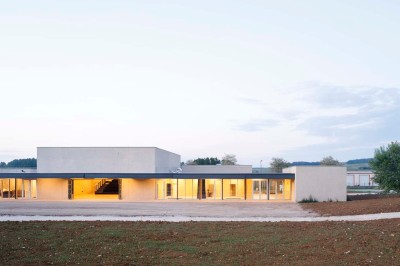 Paysage rural avec des champs cultivés, une maison isolée et une forêt en arrière-plan sous un ciel bleu.