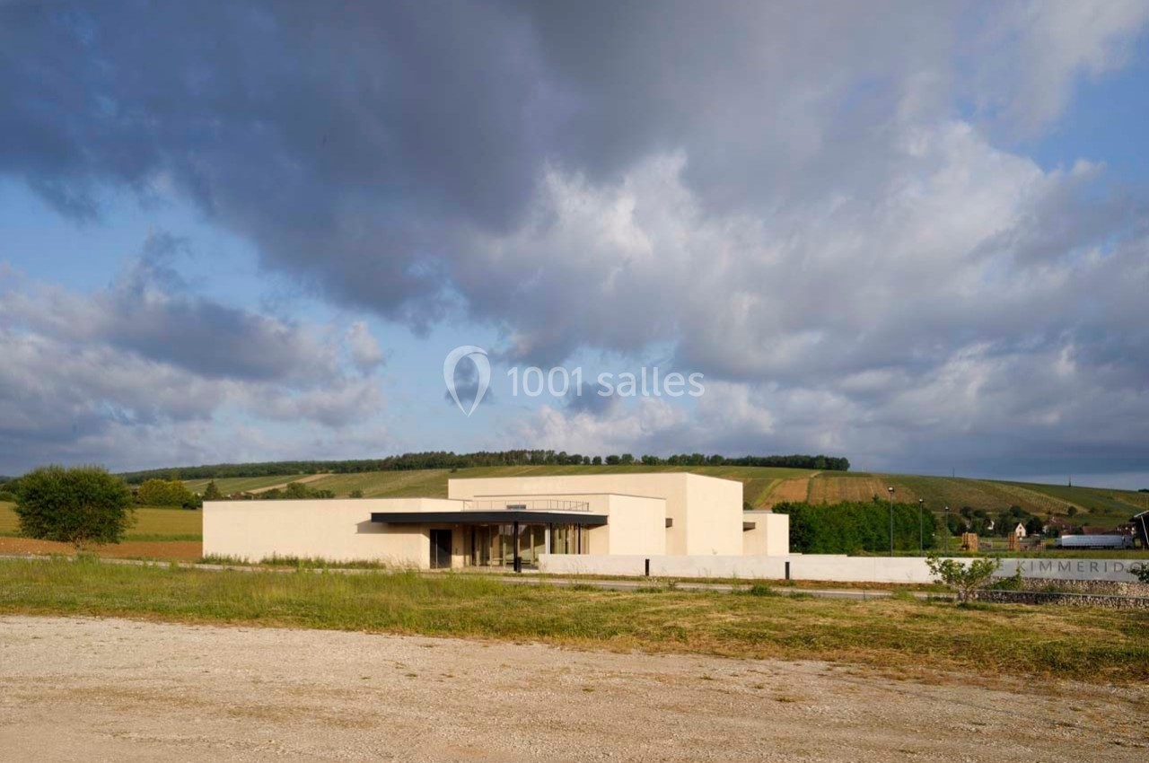 Bâtiment moderne beige entouré de champs et de collines sous un ciel partiellement nuageux.