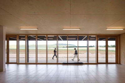 Paysage rural avec des champs cultivés, une maison isolée et une forêt en arrière-plan sous un ciel bleu.