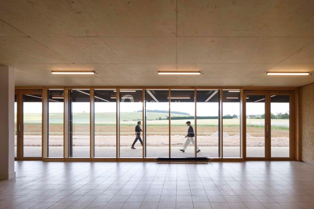 Deux personnes marchent à l'extérieur devant une large baie vitrée donnant sur un paysage de champs et collines.