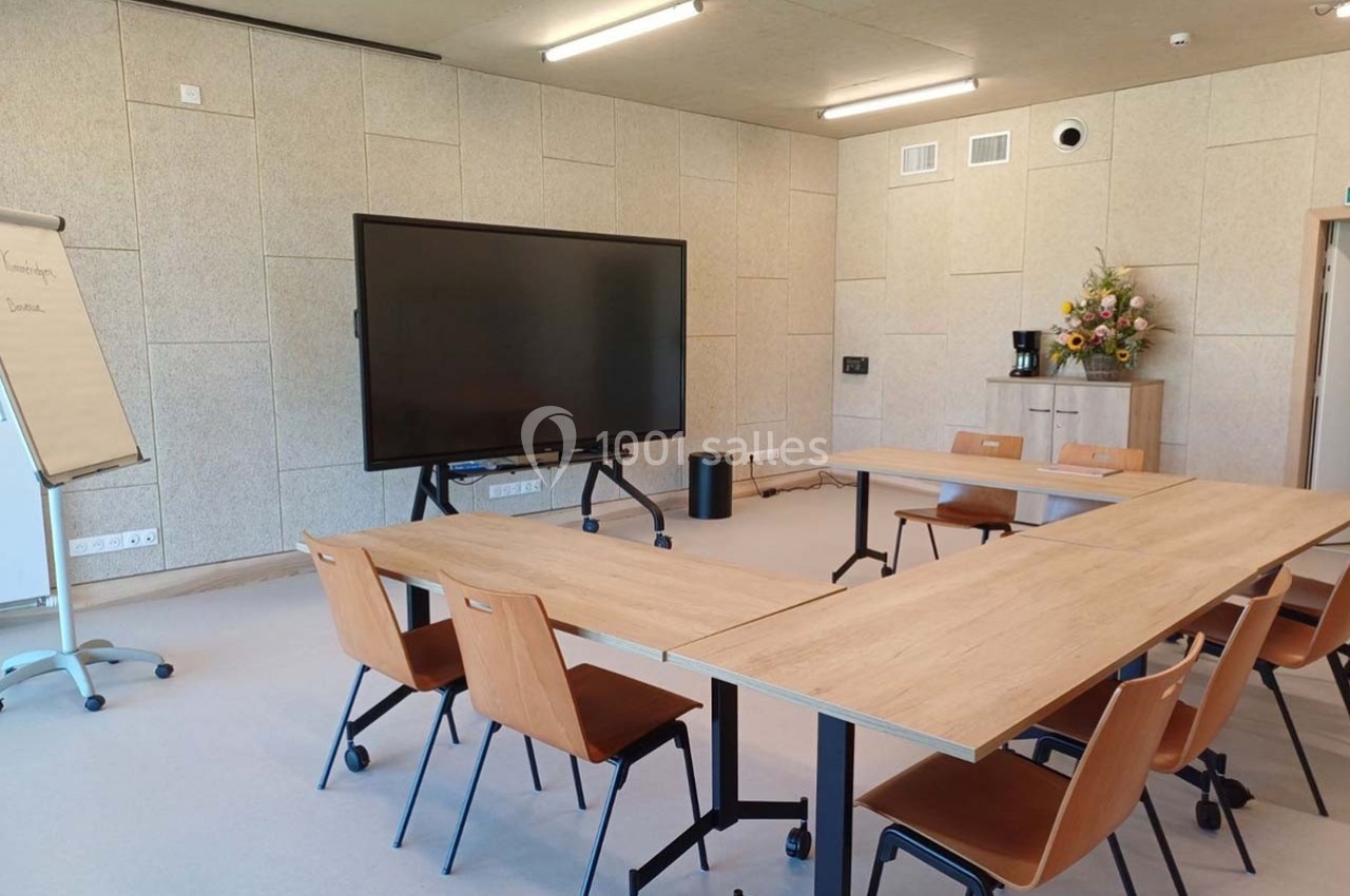 Salle de réunion lumineuse avec tables en bois, chaises, écran interactif, tableau papier et bouquet de fleurs.