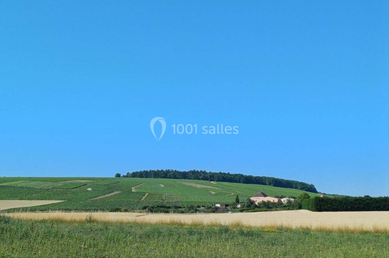 Paysage rural avec des champs cultivés, une maison isolée et une forêt en arrière-plan sous un ciel bleu.