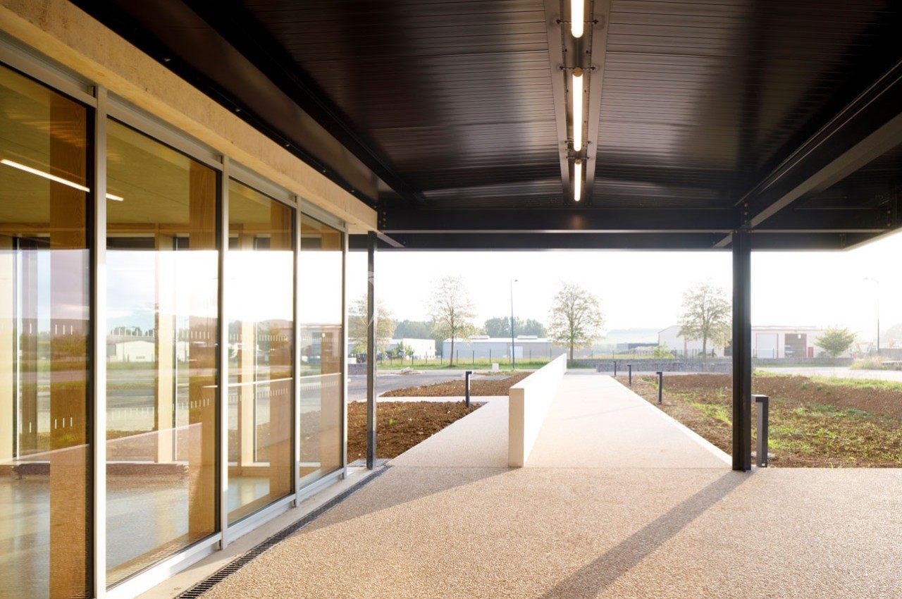 Passage couvert moderne avec structure métallique, grandes baies vitrées et vue sur un espace extérieur dégagé.