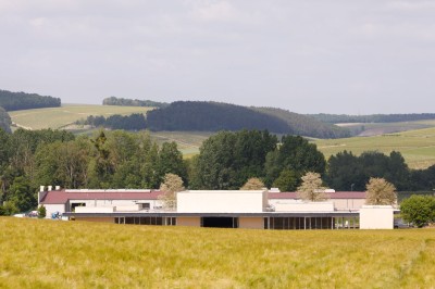 Paysage rural avec des champs cultivés, une maison isolée et une forêt en arrière-plan sous un ciel bleu.