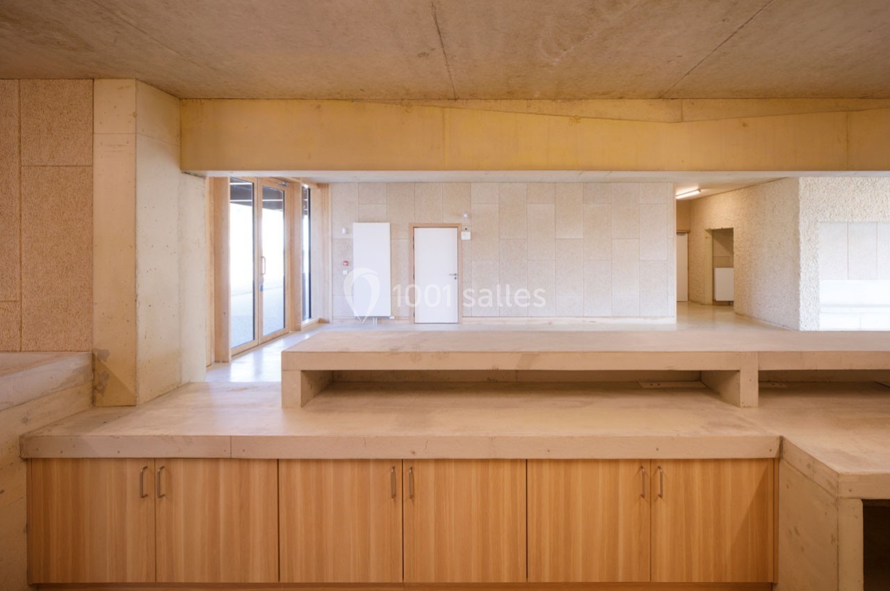 Intérieur d'un bâtiment avec murs en béton, mobilier en bois clair et lumière naturelle entrant par une baie vitrée.