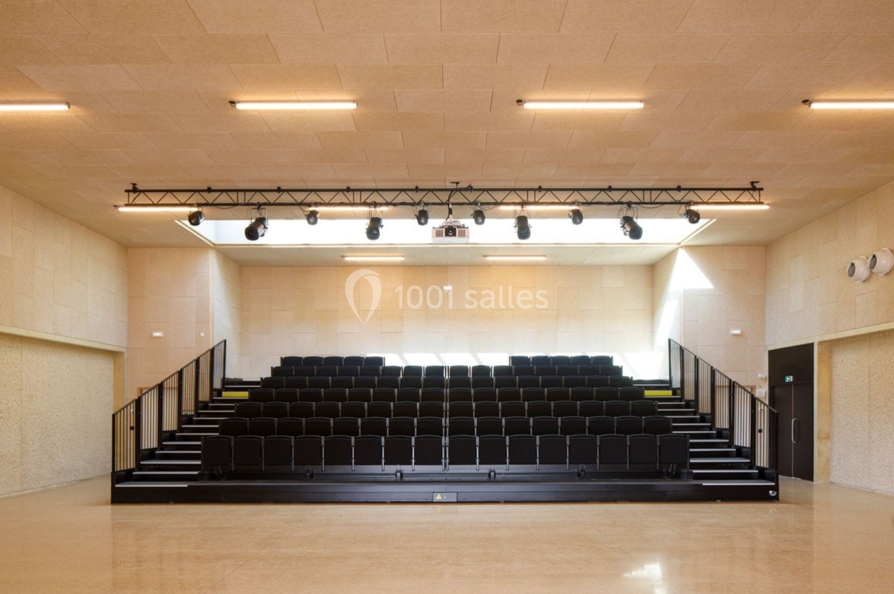 Gradins noirs vides dans une salle lumineuse avec un sol beige, des murs clairs et un éclairage suspendu.