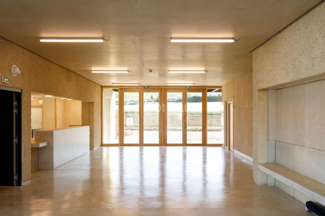 Salle intérieure lumineuse avec sol en béton poli, murs clairs, grandes baies vitrées et vue sur un paysage extérieur.