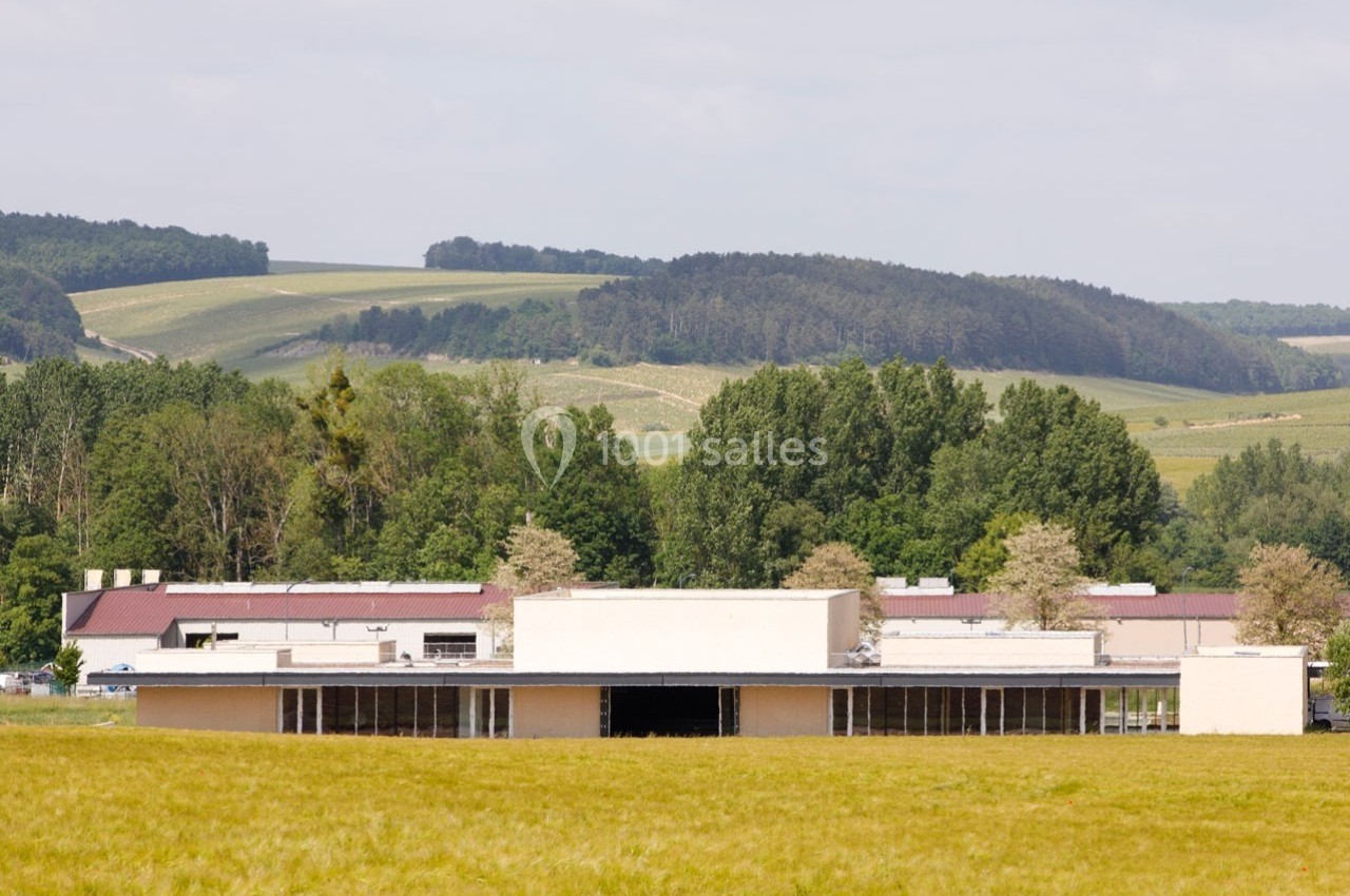 Bâtiment moderne entouré de champs et d'arbres, avec des collines boisées en arrière-plan sous un ciel clair.