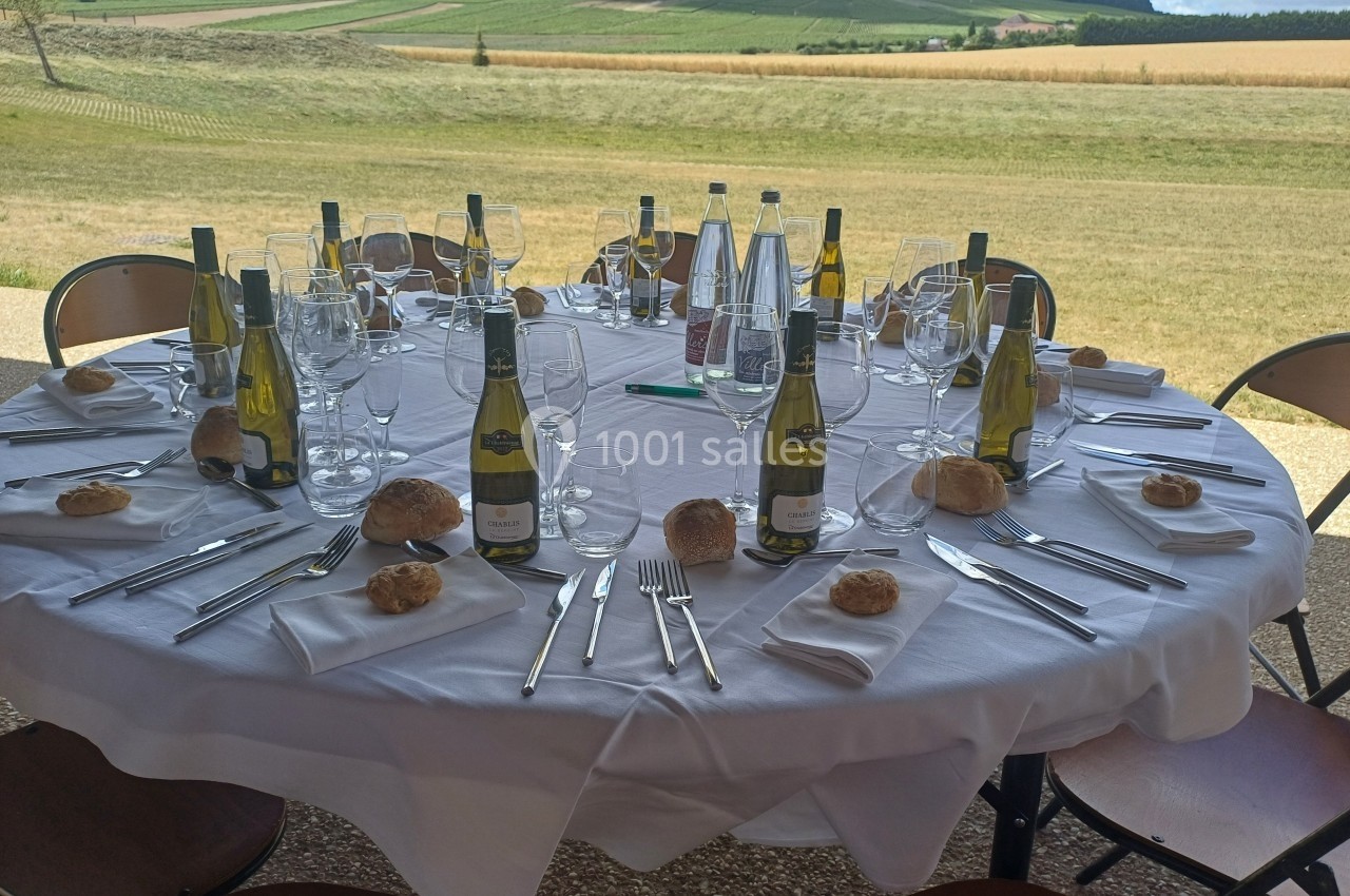 Table ronde dressée en extérieur avec nappes blanches, couverts, verres, bouteilles de vin et pains individuels.