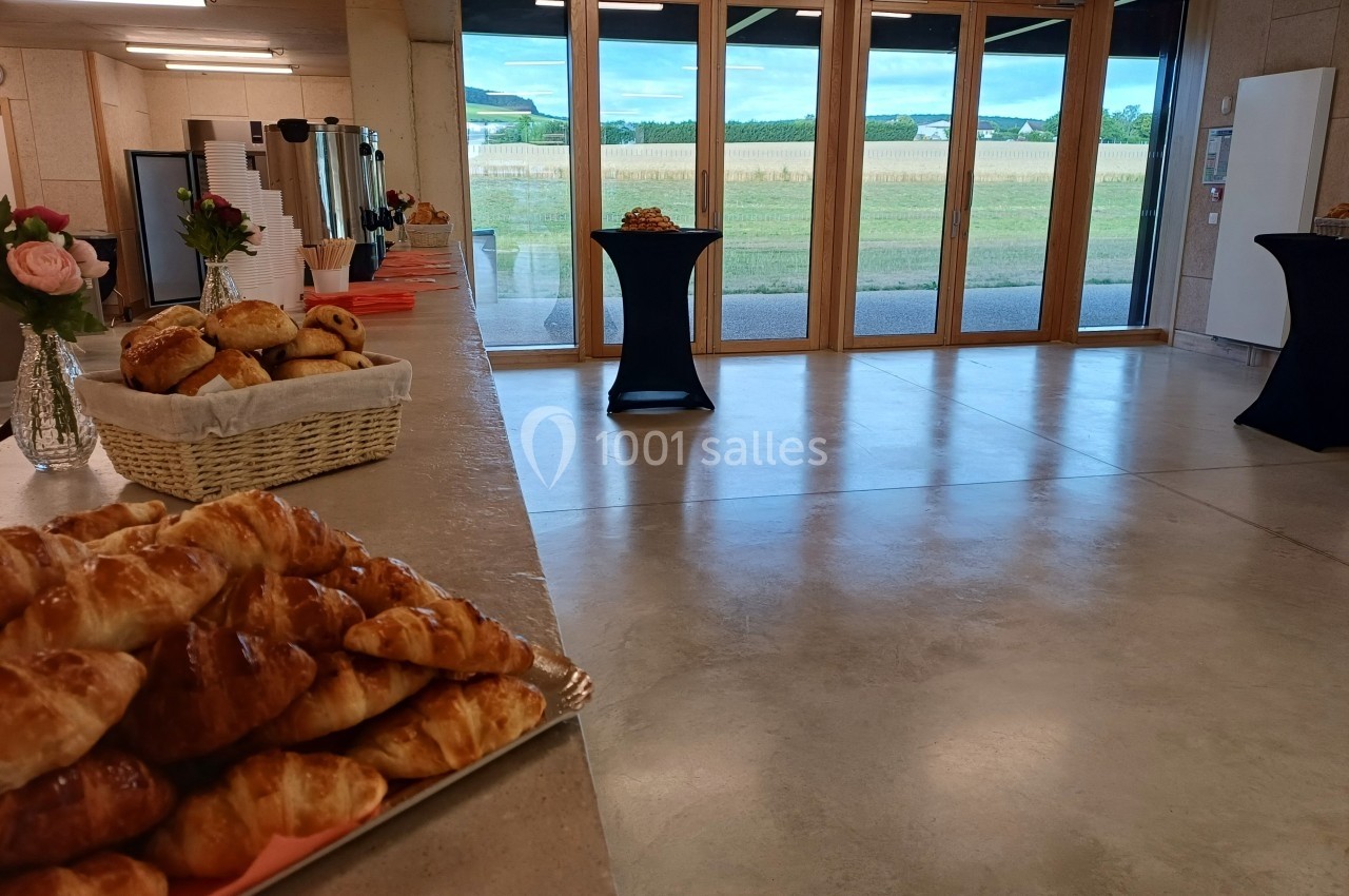 Salle lumineuse avec baies vitrées donnant sur un paysage champêtre, tables hautes et viennoiseries en premier plan.